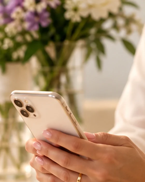 Hands holding a phone beside a vase of white and purple flowers